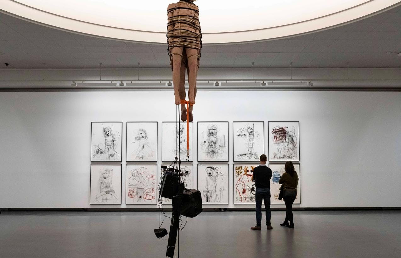 Fotografi fra inne i utstillingen til Paul McCarthy hvor vi ser voksfigurer av en naken kvinne og en naken mann som ligner Hitler som henger fra taket med høyttalere festet til bena. I bakgrunnen ses 12 tegninger i ramme på veggen. To mennesker ser på tegningene. 