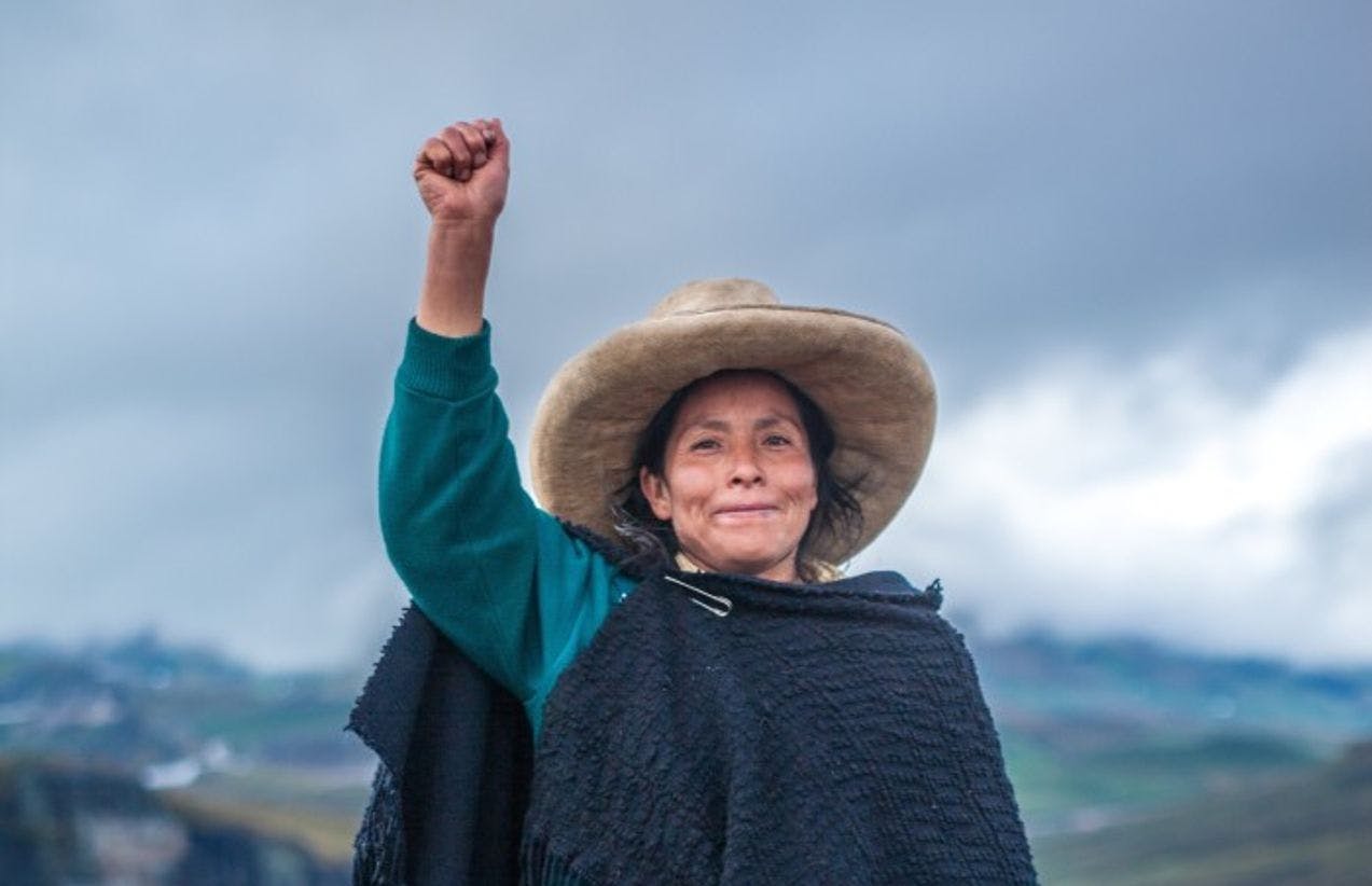 Et fotografi av en kvinne i tradisjonelle klær fra Peru som står med en knyttet hånd løftet, og et lett smil.